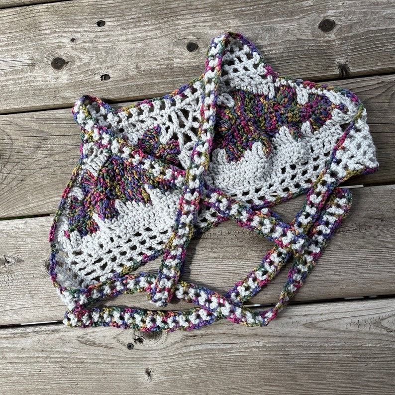 Puede incluir: Top de crochet blanco hecho a mano con un dise&ntilde;o floral en el centro. El top presenta un cuello halter y correas anudadas, con un patr&oacute;n colorido y multicolor en el dise&ntilde;o floral. El crochet es abierto y aireado.