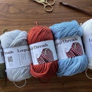 Op de afbeelding: Vijf bollen Loops & Threads Luxe Merino garen in olijfgroen, lichtgrijs, roestoranje, lichtblauw en wit. Elke bol heeft een wit papieren label met merknaam en QR-code. Een houten haaknaald en een gouden schaar zijn ook in beeld zichtbaar.