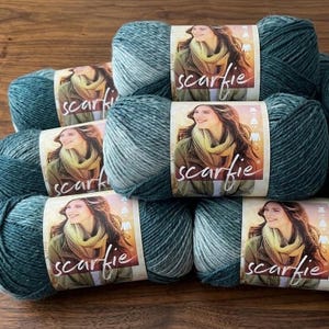 May include: Several skeins of teal and light blue variegated yarn are stacked. Each skein has a label with the word "scarfie" and a woman wearing a scarf. The yarn is arranged on a wooden surface.