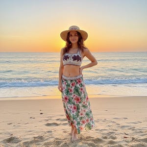 Puede incluir: Una mujer con un top de ganchillo, una falda floral y un sombrero de paja est&aacute; de pie en una playa de arena al atardecer. El top es de color crema con un dise&ntilde;o floral oscuro. La falda presenta un estampado floral tropical con flores rosas y hojas verdes.