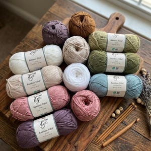 May include: A collection of Yarn Bee Bamboo-Tiful yarn in various colors, including white, beige, pink, purple, and green, arranged on a wooden surface. The image also features wooden knitting needles and a few wooden beads.
