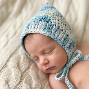 May include: A hand-crocheted baby bonnet in shades of blue and white, featuring a pointed crown and tie fastenings. The bonnet is made from soft yarn and is placed on a cream cable-knit blanket. The colour scheme includes blue, white, and a hint of green.