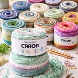 Op de afbeelding: Een verzameling Caron Skinny Cakes garen in verschillende kleuren, waaronder roze, blauw, groen en geel. De afbeelding toont meerdere garen cakes, sommige met labels, samen met breinaalden, scharen en steekmarkeerders. Het garen is gerangschikt op een lichtgekleurd houten oppervlak.