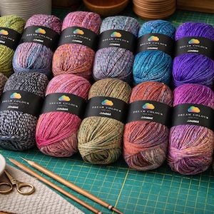 Peut inclure: Seize écheveaux de laine multicolore disposés en deux rangées, chacun entouré d'une bande de papier noir marquée Dream Colour par Hobbii. Les fils présentent des tons de vert, bleu, violet, rose, orange et marron. Deux aiguilles à tricoter en bois, un petit bol de perles colorées et des ciseaux dorés sont placés au premier plan.