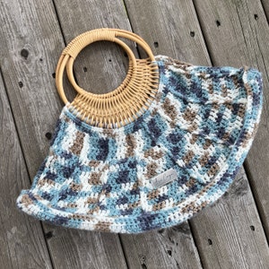 Könnte beinhalten: Eine handgefertigte Häkeltasche mit einem gewebten Rattangriff. Die Tasche hat ein geometrisches Muster in Blau-, Weiß- und Brauntönen. Ein kleines silbernes Etikett mit dem Text "Annie Cooper" ist angebracht.