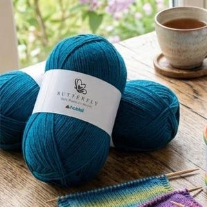 May include: Three teal yarn balls with white labels that read "BUTTERFLY" and "hobbii". A cup of tea sits on a wooden coaster. Knitting needles and a partially knitted piece are in the foreground. The yarn is 100% premium acrylic.