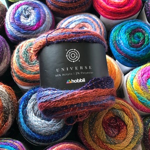 Könnte beinhalten: Nahaufnahme eines Garnknäuels mit einem mehrfarbigen, glitzernden Design. Das Garn ist mit "Universe" beschriftet und besteht aus 98% Acryl und 2% Polyester. Das Etikett enthält auch den Markennamen "hobbii".