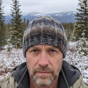 Może przedstawiać: Prążkowana czapka z dzianiny w odcieniach szarości, brązu i bieli. Czapka jest noszona przez osobę w szarej i oliwkowej kurtce, na tle ośnieżonych gór.