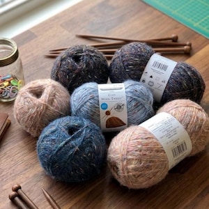 Könnte beinhalten: Eine Ansammlung von Wollknäueln in verschiedenen Farben, darunter Blau, Braun und gesprenkeltes Grau, auf einer Holzoberfläche angeordnet. Stricknadeln und ein Glas mit Maschenmarkierern sind ebenfalls zu sehen. Die Wollknäuel tragen das Etikett "Cozy Wool Tweed".
