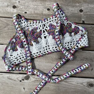 Puede incluir: Un top de ganchillo blanco con un dise&ntilde;o floral y acentos coloridos. El top presenta un cuello halter y correas para atar. El trabajo de ganchillo es intrincado, con una mezcla de patrones s&oacute;lidos y calados. Los acentos coloridos incluyen morado, rosa y verde.