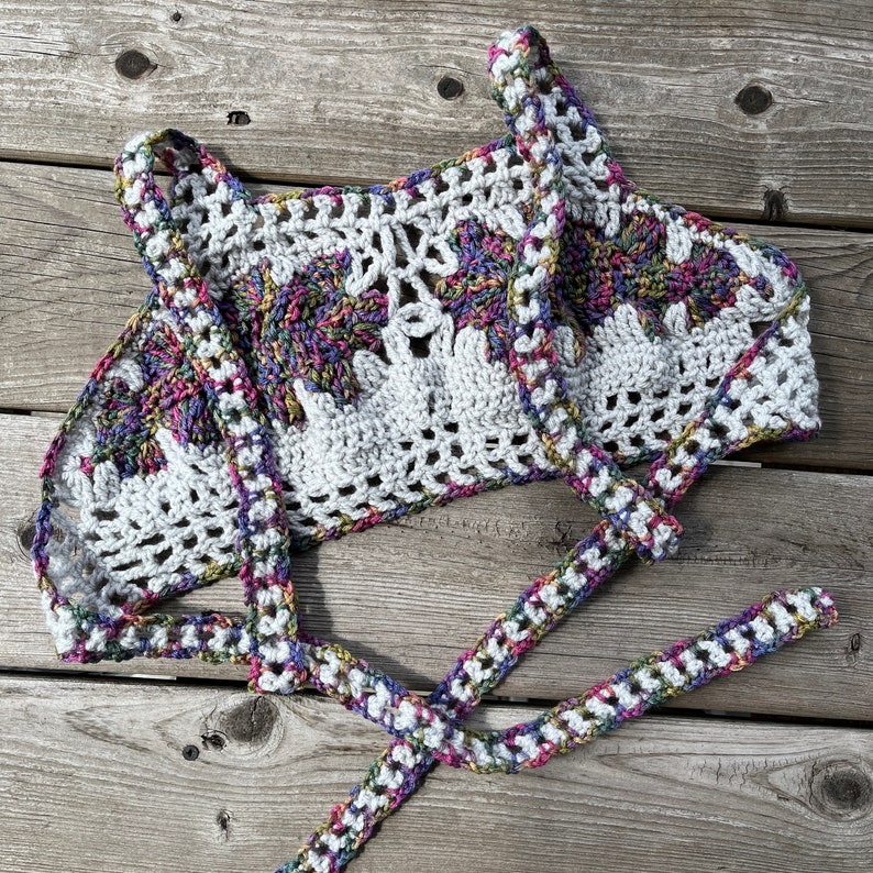 Puede incluir: Un top de ganchillo blanco con un dise&ntilde;o floral y acentos coloridos. El top presenta un cuello halter y correas para atar. El trabajo de ganchillo es intrincado, con una mezcla de patrones s&oacute;lidos y calados. Los acentos coloridos incluyen morado, rosa y verde.