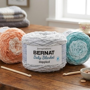 May include: Three skeins of Bernat Baby Blanket dappled yarn in orange, gray, and teal. A wooden crochet hook rests near the gray yarn. A small bowl of wooden buttons and a teacup are in the background.