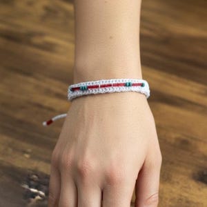 Könnte beinhalten: Ein weißes Häkelarmband mit roten und türkisfarbenen Akzenten. Das Armband ist mit kleinen türkisfarbenen Perlen und einer roten Mittellinie verziert. Das Armband befindet sich an einem Handgelenk, vor einem Holzgrund.