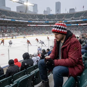 May include: A person wearing a red jacket, blue jeans, and a black, red, and white knit hat watches a hockey game in a snowy stadium. The person is holding a cup of coffee. The hockey game is in progress on the ice rink.