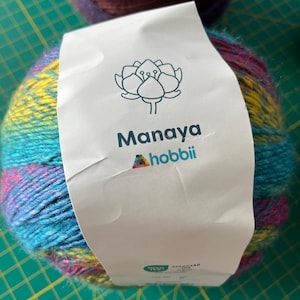 Op de afbeelding: Een bol kleurrijk garen met een label met de tekst "Manaya hobbii". Het garen is een mix van blauw, geel, roze en paars. Het label heeft een bloemenontwerp.