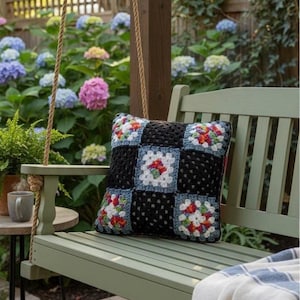 May include: A black crochet pillow with colorful floral squares rests on a green wooden swing. The swing is on a wooden deck, with a blue and white striped blanket draped over the seat. Hydrangeas and other greenery are in the background.