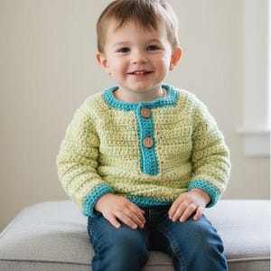 May include: A child's handmade crochet sweater in light green with turquoise trim and wooden buttons. The sweater is paired with blue jeans and white socks. The child is smiling.