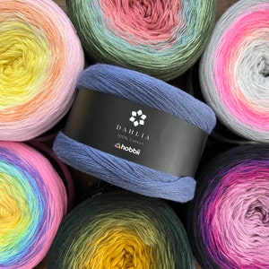 May include: A collection of colorful yarn skeins, including a blue skein with a black label that reads "DAHLIA 100% Cotton" and the brand "hobbii". The other skeins display a gradient of colors, such as pink, yellow, green, and purple.