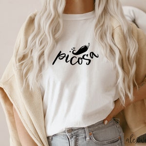 May include: White t-shirt with a black graphic of a chili pepper and the word "picosa" in a handwritten font.
