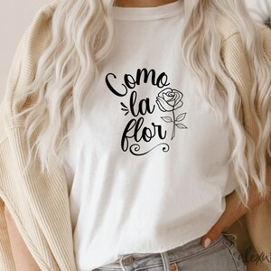 May include: White t-shirt with the text "Como la flor" in black cursive lettering and a black outline drawing of a rose.