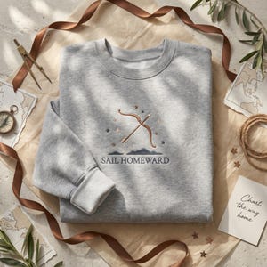 May include: A light grey crewneck sweatshirt featuring an embroidered design of a bow and arrow surrounded by small stars and a mountain silhouette. Below the graphic, the text reads SAIL HOMEWARD in a dark blue, capitalised, sans-serif font.
