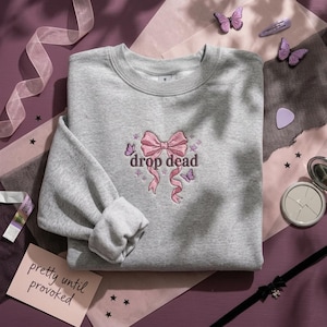 May include: A heather gray crewneck sweatshirt featuring an embroidered design of a pink ribbon bow, small purple butterflies, and stars, with the text "drop dead" in dark brown lettering centered on the chest of the garment.
