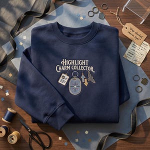 Op de afbeelding: Een marineblauw sweatshirt met ronde hals en geborduurde tekst "HIGHLIGHT CHARM COLLECTOR" boven een verzameling kleine bedeltjes, waaronder een rechthoekig label, een ovaal hangertje met een ster, twee pijlen en een vogel-silhouet.