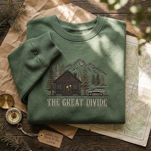 May include: A sage green crewneck sweatshirt featuring an embroidered design of a cabin in the woods with mountains, pine trees, and a small vehicle. The text THE GREAT DIVIDE is embroidered below the cabin in a serif font.