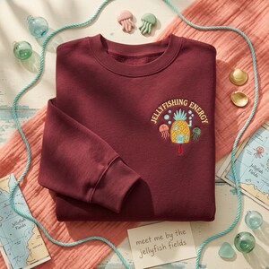 May include: A maroon long-sleeve sweatshirt featuring an embroidered design on the upper left chest. The embroidery depicts a yellow pineapple house, two small jellyfish, and the text "JELLYFISHING ENERGY" in a curved yellow font.
