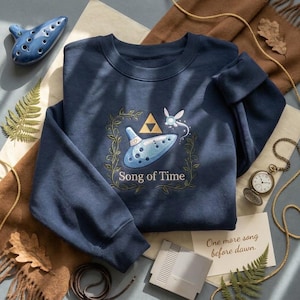 Peut inclure: Un sweat-shirt bleu marine à col rond orné d'une broderie représentant un ocarina bleu clair, un symbole Triforce doré et une petite fée. Le texte "Song of Time" est brodé sous l'ocarina, entouré d'une bordure de vigne verte délicate.