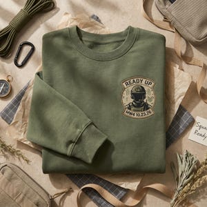 May include: An olive green crewneck sweatshirt featuring a circular patch on the left chest. The patch displays a soldier in tactical gear with the text READY UP and MW4 10.23.26 printed in a dark brown font against a tan background.