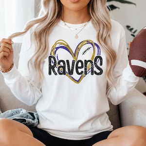 May include: A white long-sleeve sweatshirt featuring a graphic of a heart outlined in yellow and purple scribbles with the word Ravens printed in black across the center. The design includes a small football icon inside the heart shape.