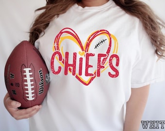 Op de afbeelding: Een wit t-shirt met een rood en geel hartmotief en het woord CHIEFS in rode, verweerde letters. Een klein geel hartje staat boven het woord en een zwarte afbeelding van veters van een football bevindt zich aan de rechterkant van het hart.