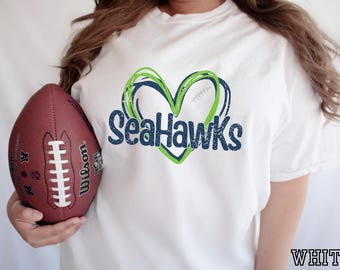 以下が含まれることがあります： ライムグリーンとネイビーブルーのハートの輪郭と、ネイビーブルーの文字でSeaHawksと書かれたグラフィックが特徴の白い半袖Tシャツ。白い靴紐が付いた茶色のレザー製アメリカンフットボールがTシャツの横に持たれています。