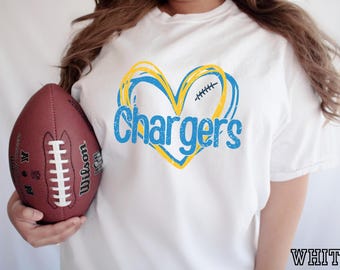 Puede incluir: Una camiseta blanca con un gráfico de un corazón delineado en amarillo y azul con la palabra Chargers en letras azules en el centro. Un balón de fútbol americano de cuero marrón con cordones blancos se sostiene al lado, mostrando el logo Wilson.