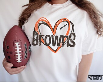May include: A white short-sleeved t-shirt featuring a graphic of a heart outlined in orange and black with a football lace detail. The word Browns is printed in a distressed dark brown font across the centre of the heart design.