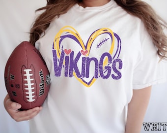 May include: A white short-sleeved t-shirt featuring a graphic of a yellow and purple heart outline with the text Vikings in purple, distressed lettering. Two small pink hearts are positioned above the text, and a brown football is held nearby.