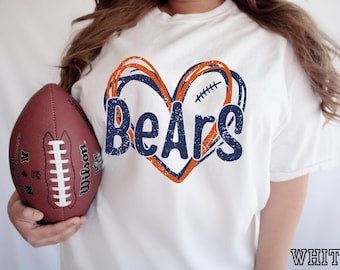 May include: A white short-sleeved t-shirt featuring a graphic of a heart outlined in orange and navy blue lines. The word "BeArs" is printed in a distressed navy blue font across the center of the heart, which also includes a small football lace detail.
