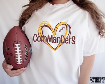 Op de afbeelding: Een wit t-shirt met een afbeelding van een hart omlijnd in geel en bordeauxrood met een detail van veters van een American football. De tekst "ComManDerS" is in een bordeauxrood, verweerd lettertype over het hart gedrukt. Een bruine football is zichtbaar.