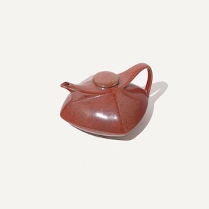 May include: A reddish-brown ceramic teapot with a lid and handle. The teapot has a unique, angular design and a small spout. The lid is circular and sits on top of the teapot. The handle is curved and attached to the side of the teapot.