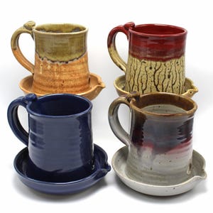 May include: Four ceramic mugs with matching saucers. The mugs are glazed in various colors, including blue, red, brown, and yellow. Each mug has a handle and a spout on the saucer.