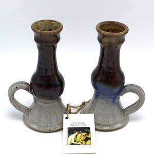 May include: A pair of handmade ceramic candle holders with handles. The holders are glazed in shades of gray, brown, and blue. The text "Eve Unity Candle Holder" is printed on a white tag.