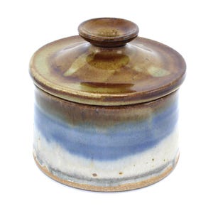 May include: A ceramic jar with a lid. The jar is cylindrical with a brown, blue, and white color scheme. The lid is brown with a small knob on top. The jar appears to be handmade and has a rustic aesthetic.