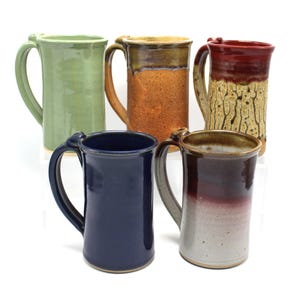 May include: Five ceramic mugs in different colors and glazes. The mugs are all the same size and shape, with a handle on the side. The mugs are arranged in a row, with the green mug on the left and the red mug on the right.