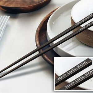 May include: A pair of black chopsticks with the text "YOUR LOGO HERE" engraved on each stick. The chopsticks are on a white plate with a brown bowl.