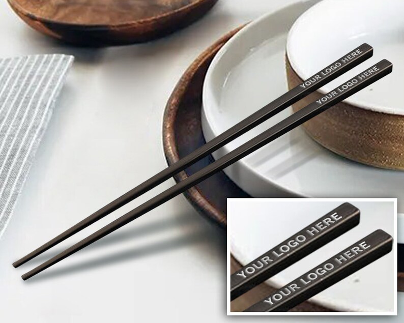 May include: A pair of black chopsticks with the text "YOUR LOGO HERE" engraved on each stick. The chopsticks are on a white plate with a brown bowl.