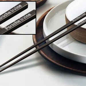 May include: A pair of black chopsticks with the words "Porsche Irvine" engraved on each chopstick. The chopsticks are resting on a white plate with a brown rim.