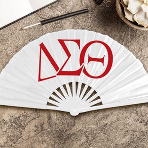 May include: A white folding fan with red Greek letters. The fan is open, displaying the letters Delta, Sigma, and Theta. The fan is on a textured, neutral-toned surface, with a pen and notebook visible in the background.