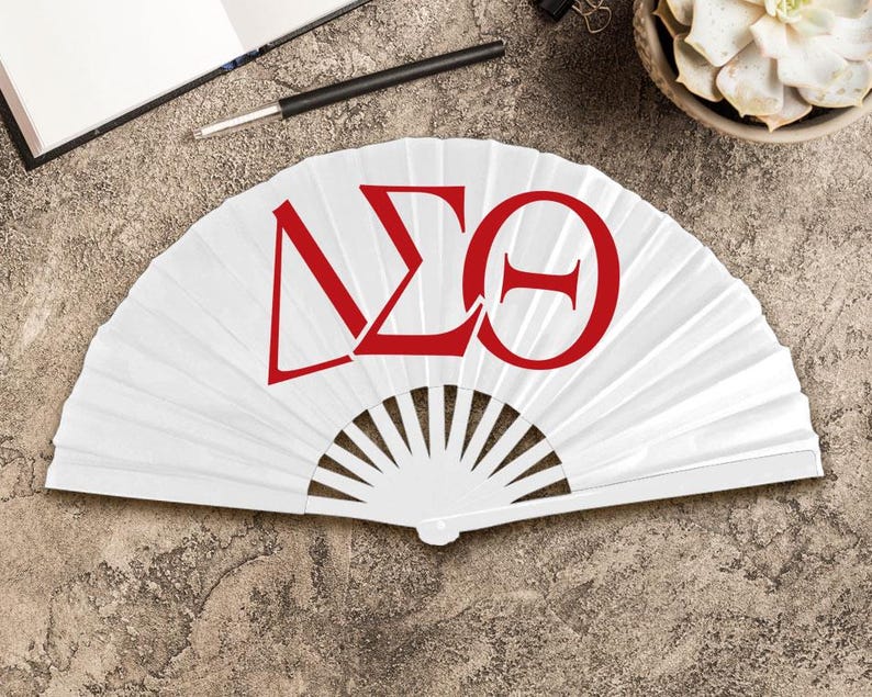 May include: A white folding fan with red Greek letters. The fan is open, displaying the letters Delta, Sigma, and Theta. The fan is on a textured, neutral-toned surface, with a pen and notebook visible in the background.