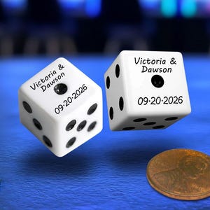 May include: Two white six-sided dice with black pips and custom black text reading Victoria & Dawson and the date 09-20-2026. The dice are suspended in the air above a blue surface next to a copper-colored penny coin.